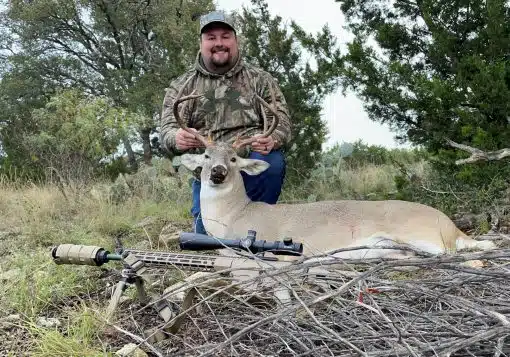 A deer hunter posing with a deer and a suppressed rifle.